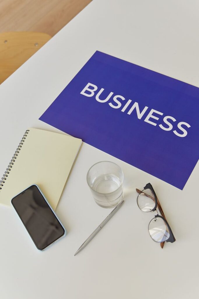 Flat lay of a desk with business-themed poster, notebook, smartphone, pen, eyeglasses, and water glass.